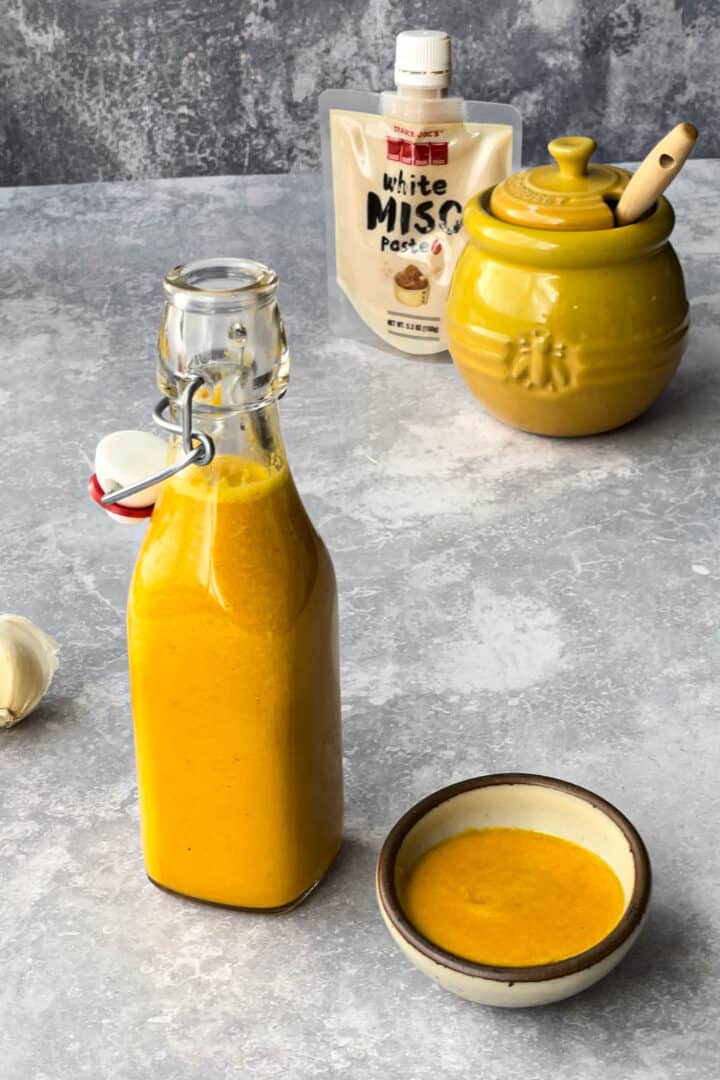 a bottle of carrot ginger miso salad dressing sits on a countertop next to a small bowl filled with the dressing.
