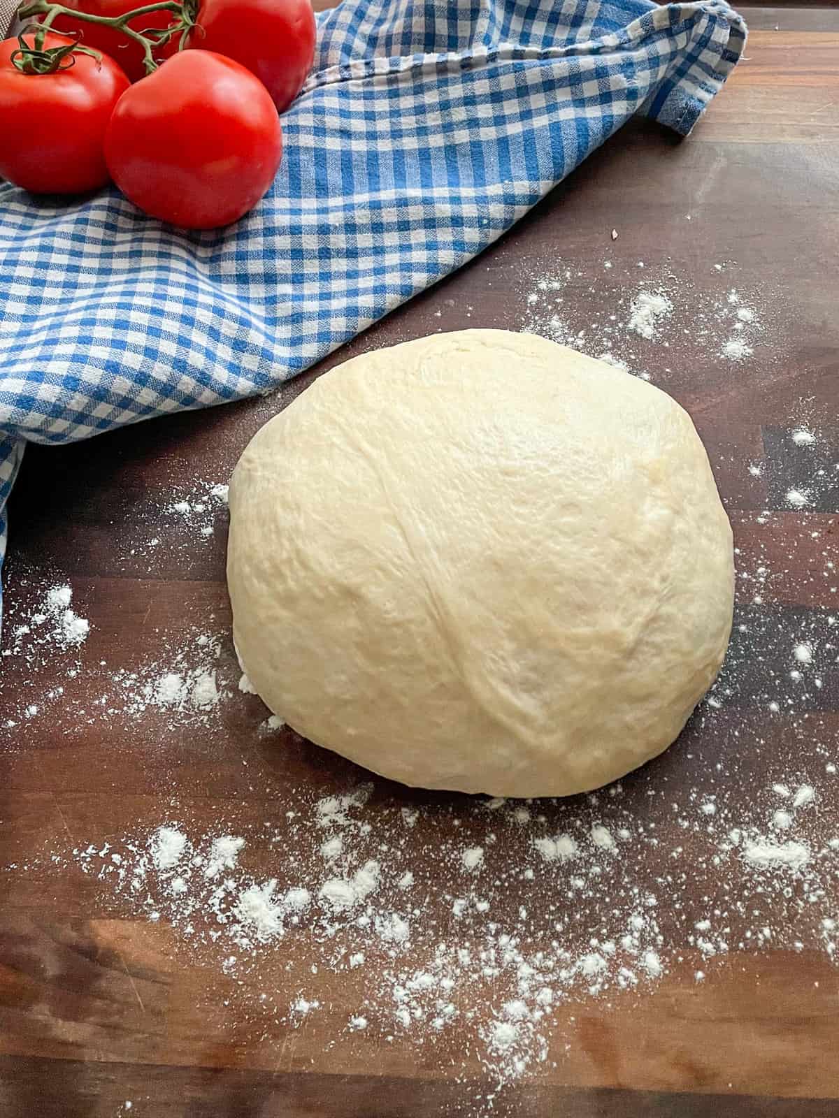 fresh homemade pizza dough sitting on top of a lightly floured cutting board next to a blue and white checkered tea towel with tomatoes on top.