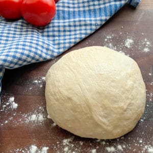 a ball of fresh, homemade pizza dough sits on a cutting board near a kitchen towel and tomatoes