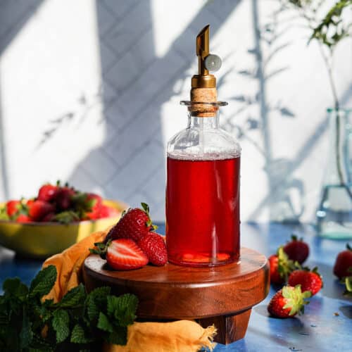 a bottle of homemade strawberry simple syrup sits on top of a wooden pedestal around ingredients used to make the recipe.