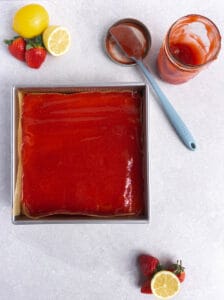 strawberry jam has been spread onto the lemon bars using a blue rubber spatula that sits on the counter.