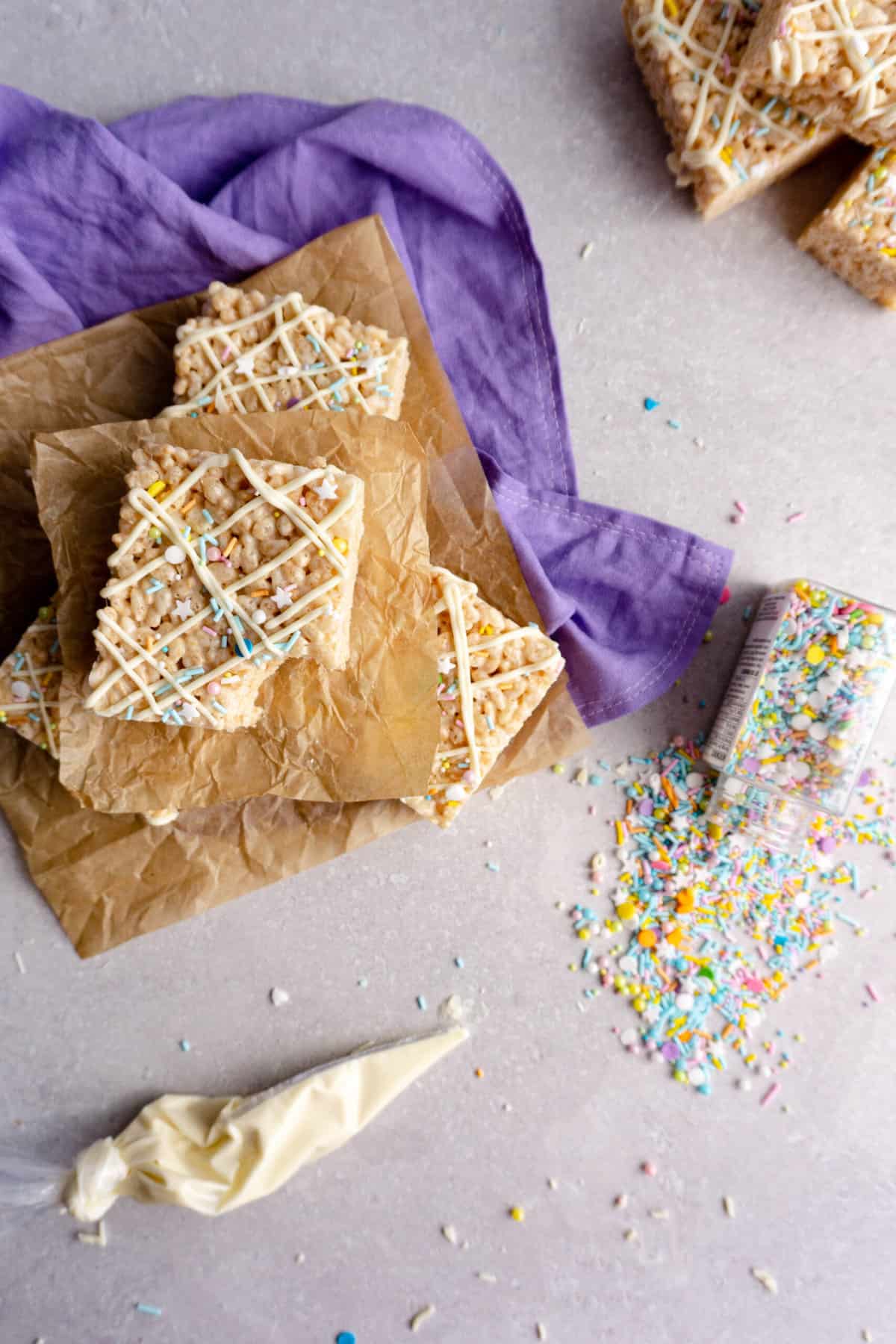 A stack of decorated copycat Disney rice krispie treats sit on top of parchment squares and a purple linen tea towel, surrounded by sprinkles and white chocolate.