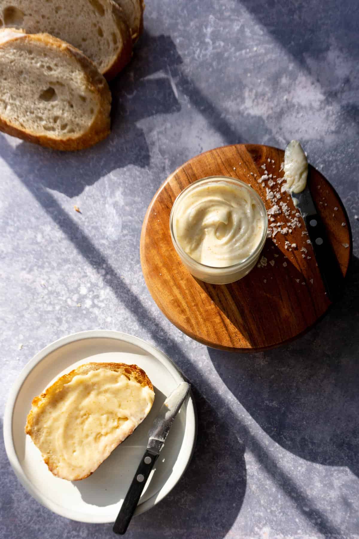 a container of whipped brown butter sits on top of a small wooden stand in the center of the photo, with a loaf of sourdough bread in the top left corner and a slice of buttered sourdough toast in the bottom left corner.