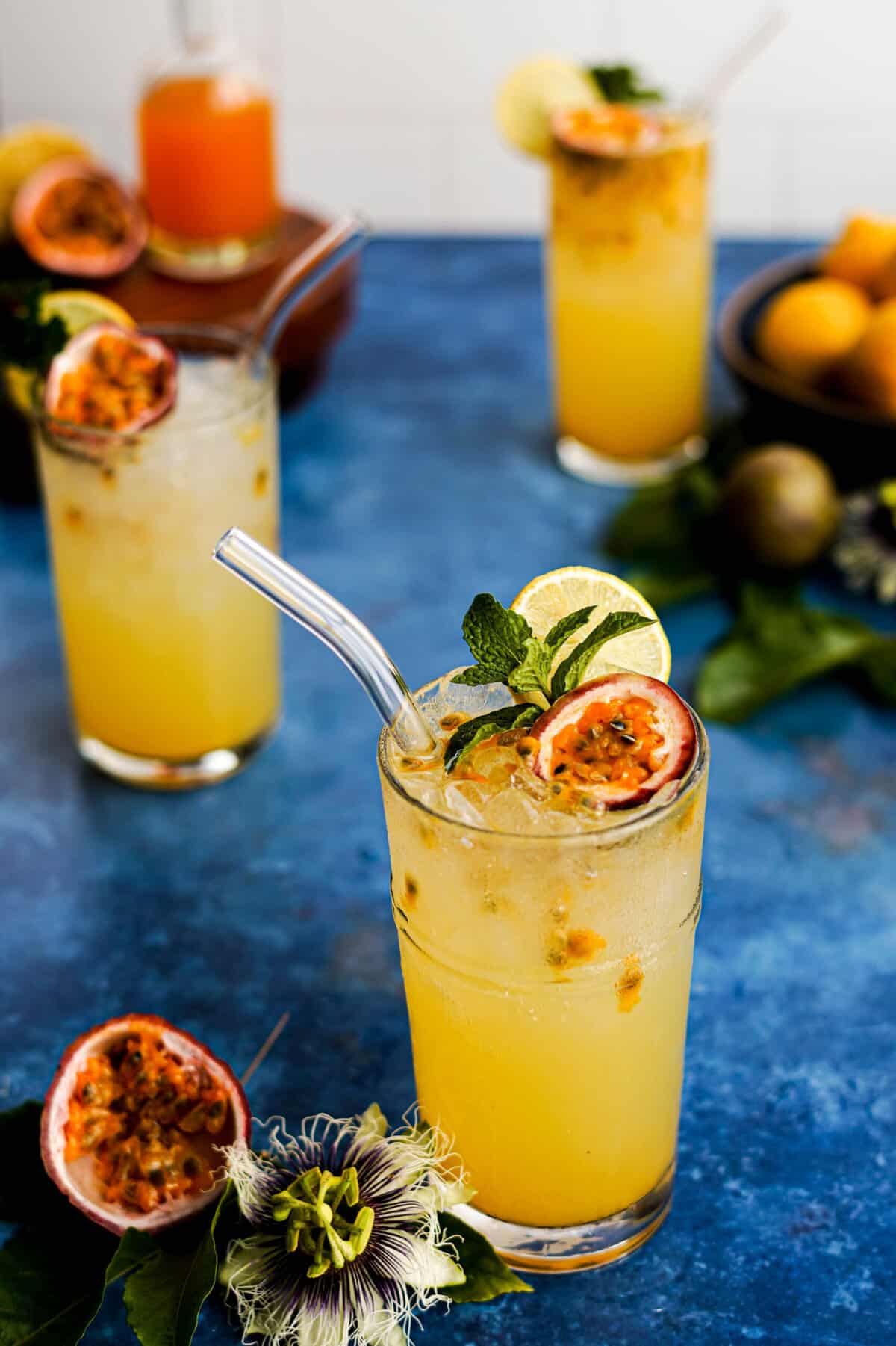 three glasses of passion fruit lemonade are on a counter top, with a small bottle of passion fruit simple syrup in the background. A sliced passion fruit sits in the foreground next to a passion flower.