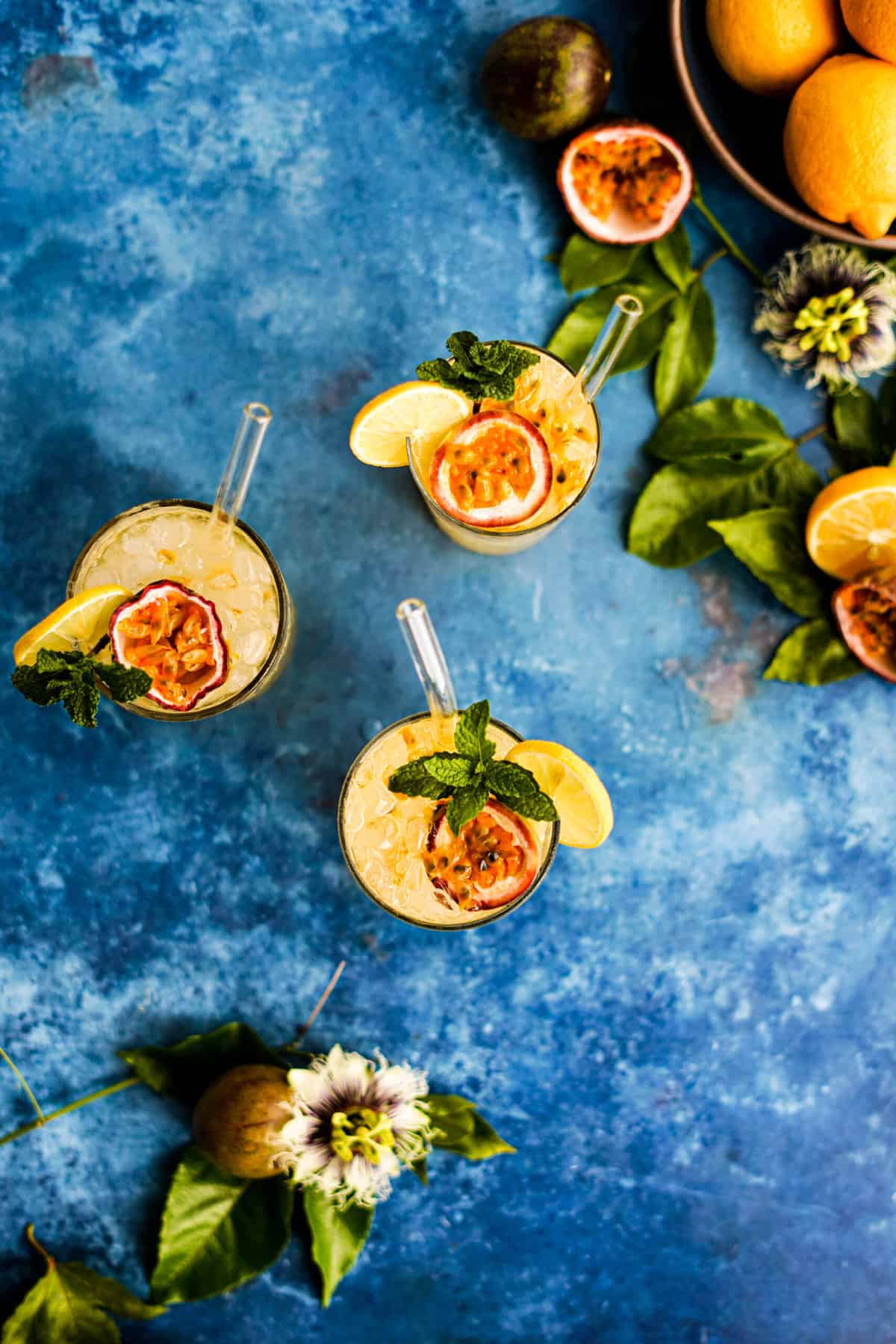 An overhead shot of three glasses of passion fruit lemonade, sitting on a table with a bowl of lemons and fresh passion fruit and passion flowers.