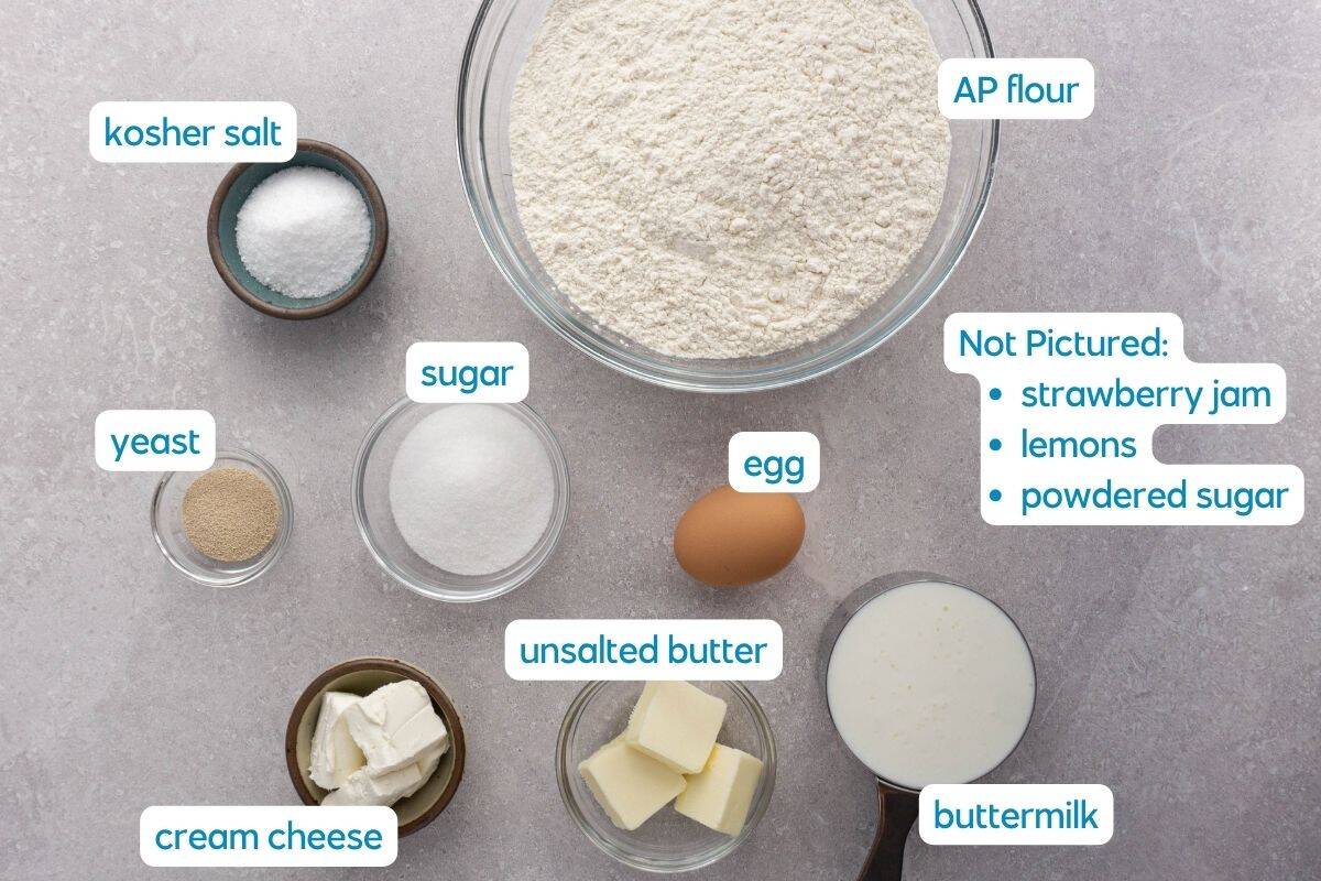A top-down view of labeled baking ingredients for strawberry cinnamon rolls: AP flour, kosher salt, yeast, sugar, cream cheese, unsalted butter, egg, and buttermilk. A note lists missing items: strawberry jam, lemons, and powdered sugar.