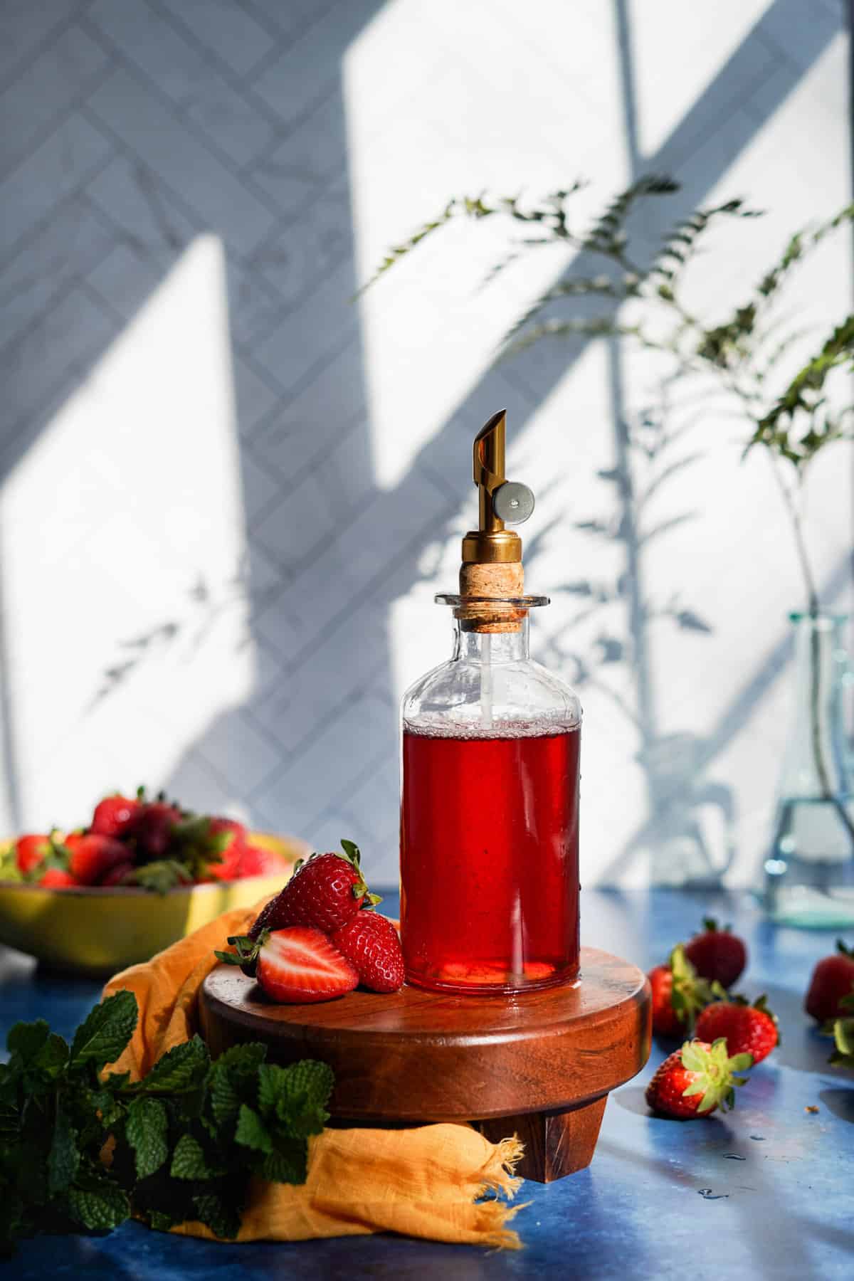 A bottle of strawberry simple syrup sits on a wooden pedestal on a blue countertop. A small pile of strawberries sits next to the bottle.