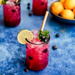 a glass of blueberry lemonade sits on a blue countertop, with a garnish of lemon slice and mint.