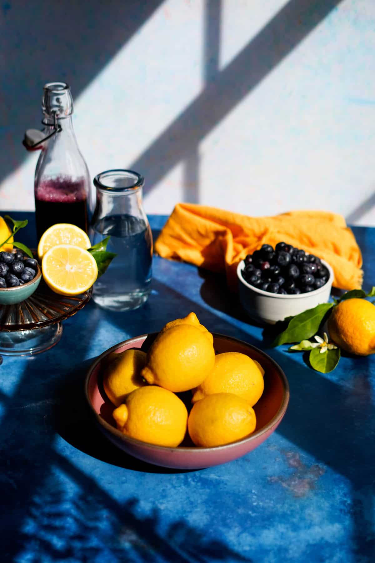 ingredients to make blueberry lemonade are sitting on a countertop, including lemons in a pink ceramic bowl, blueberries in a white ceramic bowl, and a bottle of blueberry simple syrup.