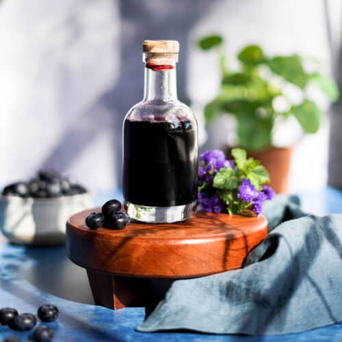 feature image of blueberry simple syrup. A bottle of homemade blueberry simple syrup sits on a small wooden pedestal.