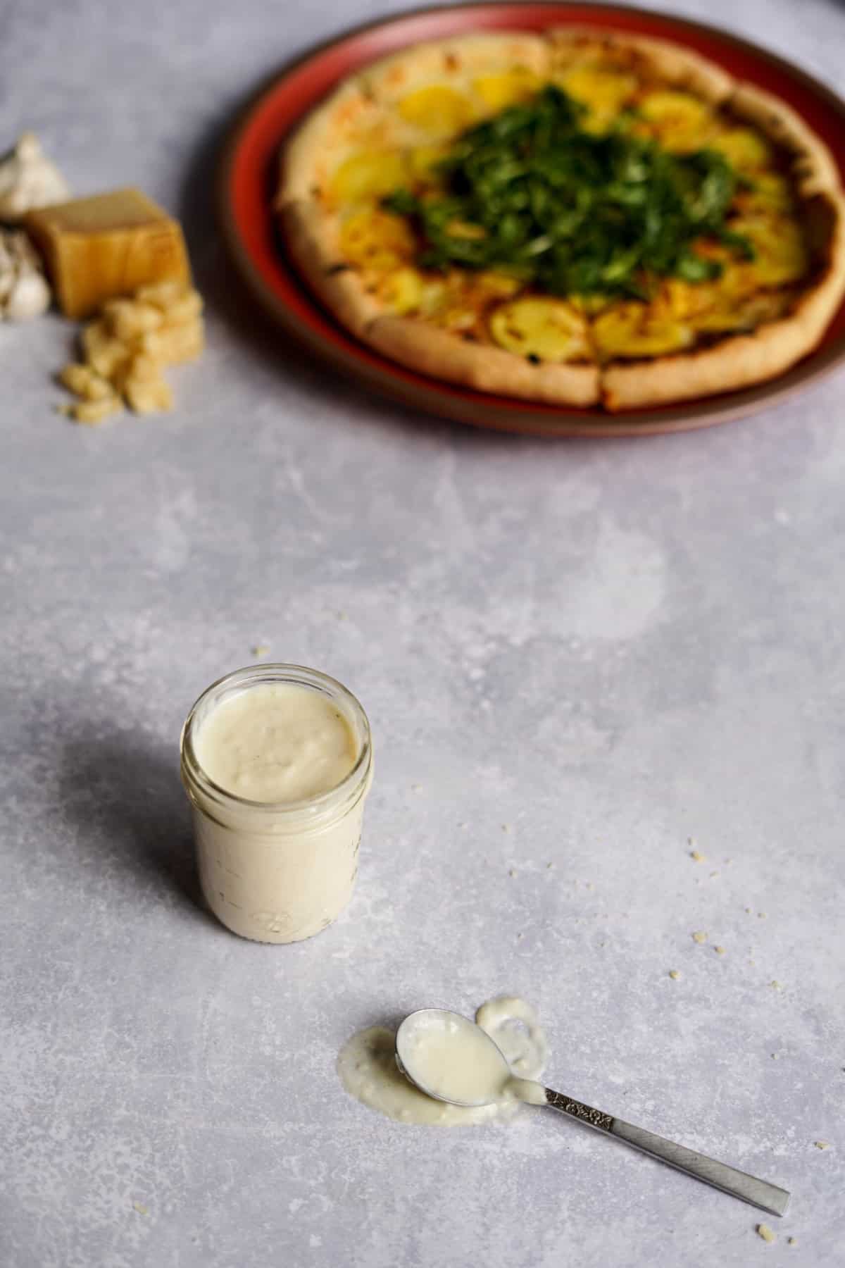 a small jar of parmesan garlic sauce sits on a counter in front of a pizza, with a spoonful of sauce to the side of the jar.