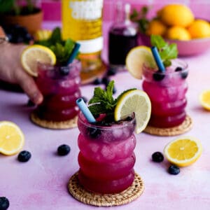 three glasses of sparkling blueberry vodka lemonades sit on a countertop, with a hand reaching to grab a glass from out of frame.