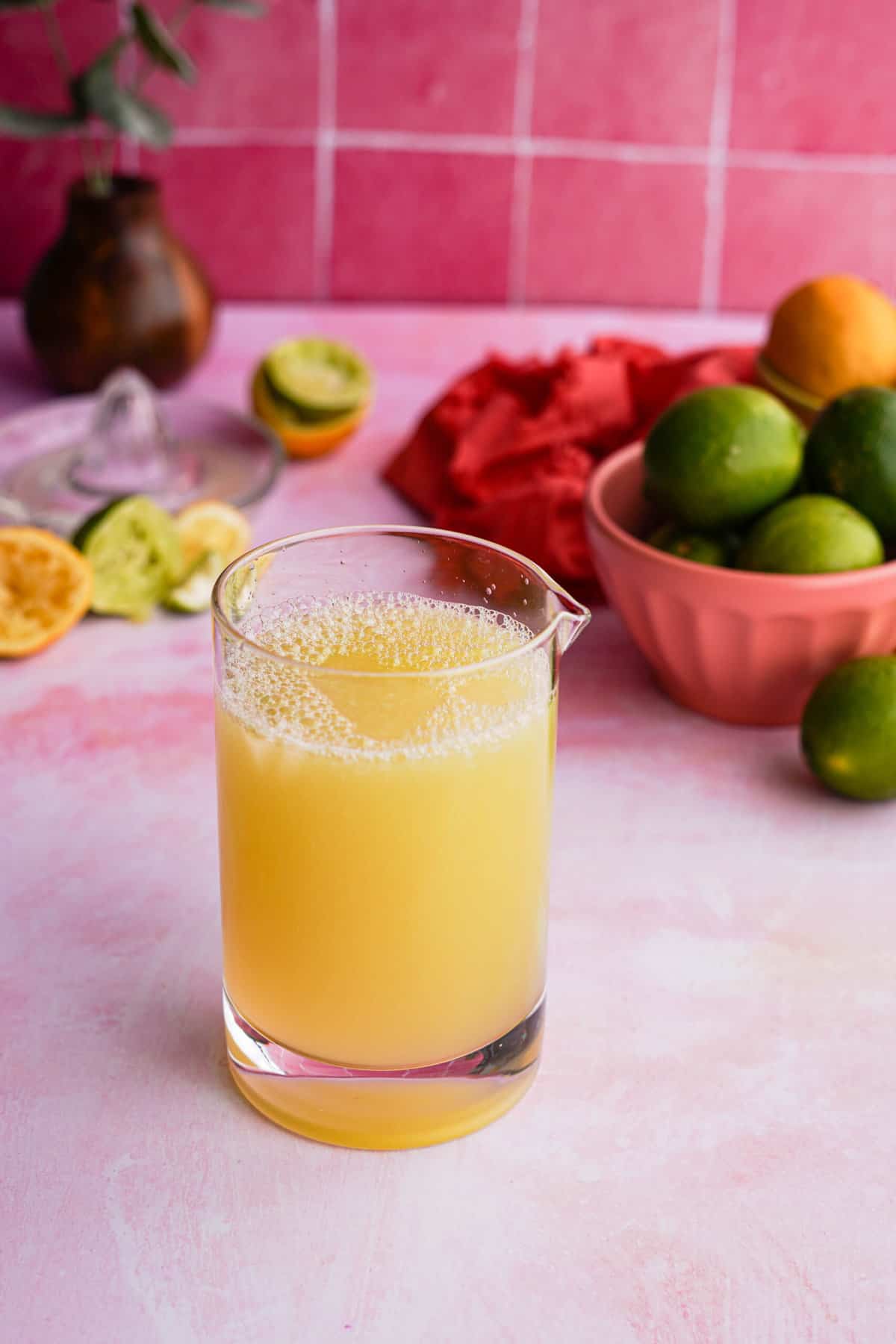 A cocktail mixing glass is full of a batch of homemade sweet and sour mix, sitting in front of bowls of lemons and limes.