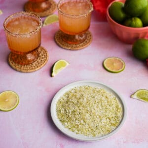 Feature image of a Lime Salt recipe, with a small dish of homemade lime salt sitting on a countertop surrounded by limes and cocktail glasses rimmed with the lime salt.