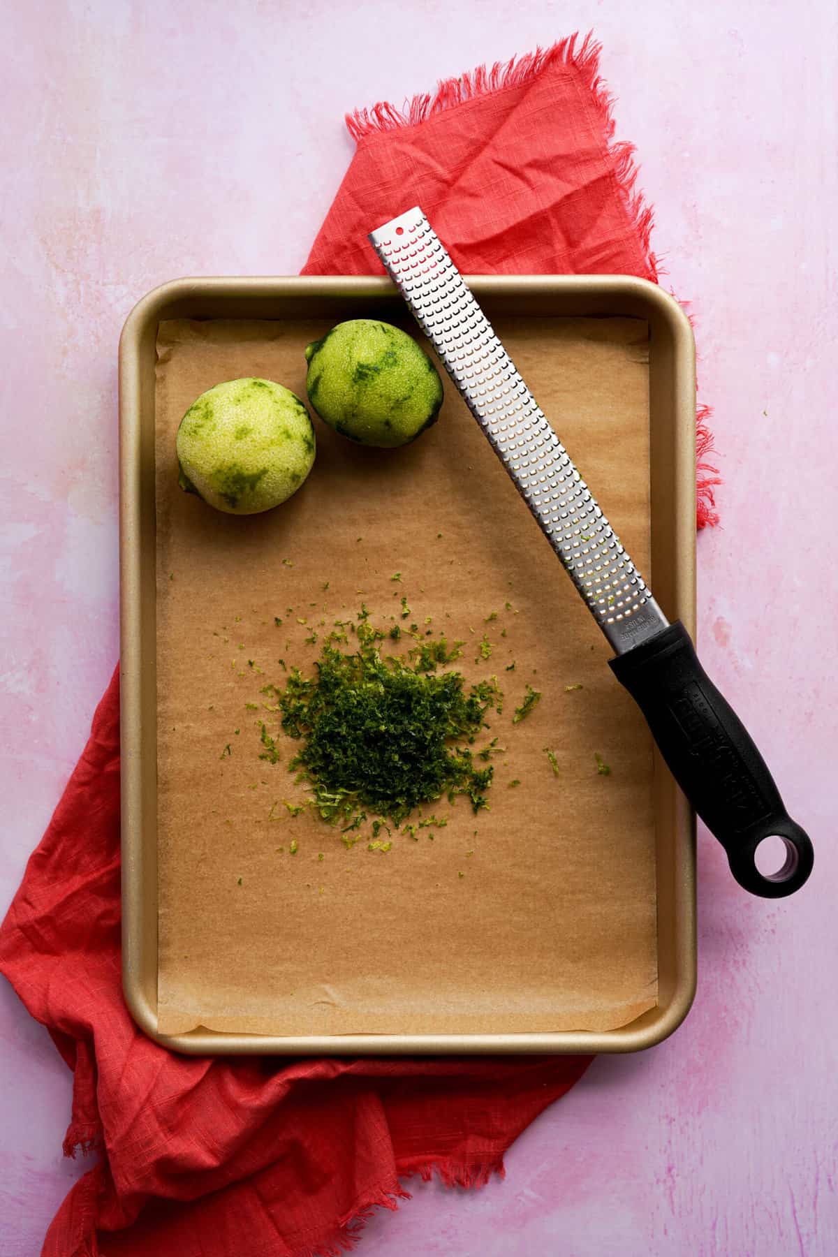 Two limes sit on a small sheet pan lined with parchment paper. They have been zested using a microplane grater and the zest of the limes sits in the center of the pan.
