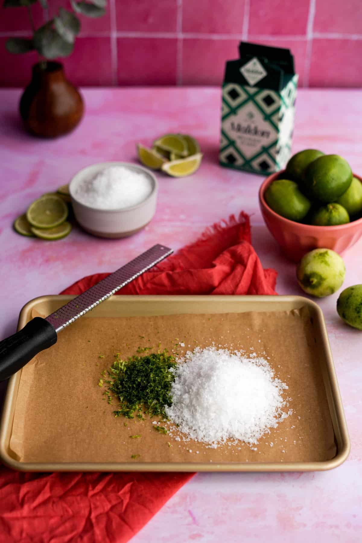 a small pile of lime zest sits next to a pile of flaky sea salt on a baking pan. A bowl of limes sits in the background of the photo, with a box of Maldon sea salt and a small dish of the salt.