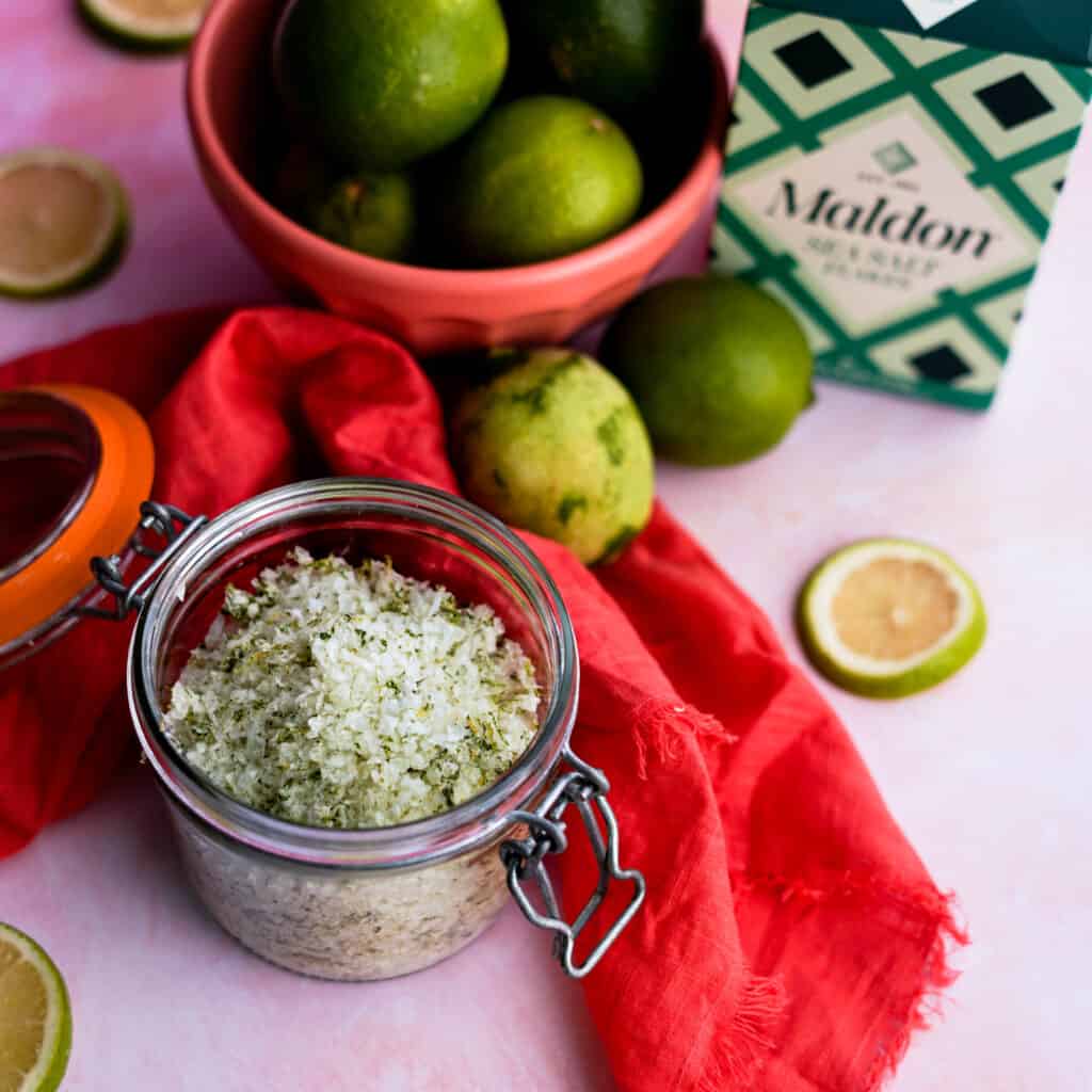 a small jar of homemade lime salt sits on a pink countertop next to a bowl of limes and a box of flaky Maldon sea salt.