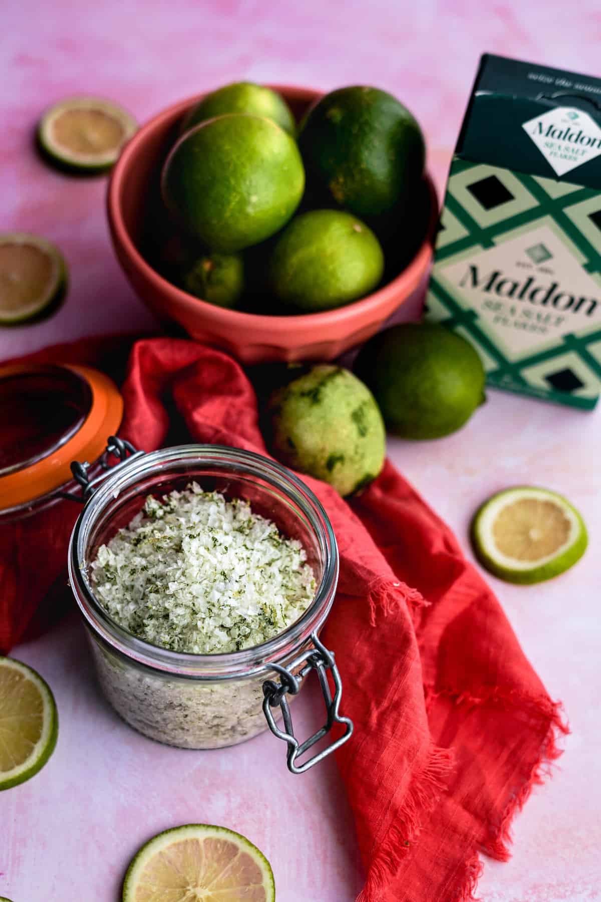 a small jar of homemade lime salt sits on a pink countertop next to a bowl of limes and a box of flaky Maldon sea salt.