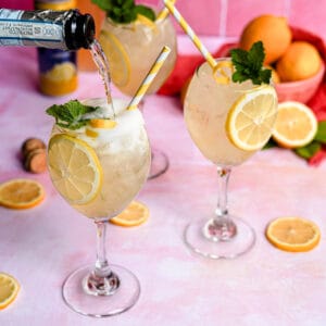 a bottle of prosecco pours into a wine glass with a limoncello spritz cocktail. Two other glasses of limoncello spritzes sit in the background, with a bowl of lemons and lemon slices.