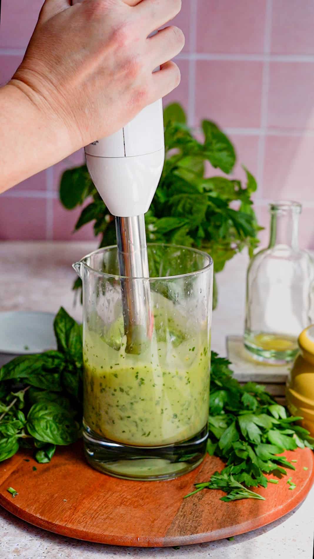 an assortment of fresh herbs (basil, chives, parsley and oregano) have been added to the base of a vinaigrette recipe, and are being mixed in using an immersion blender.