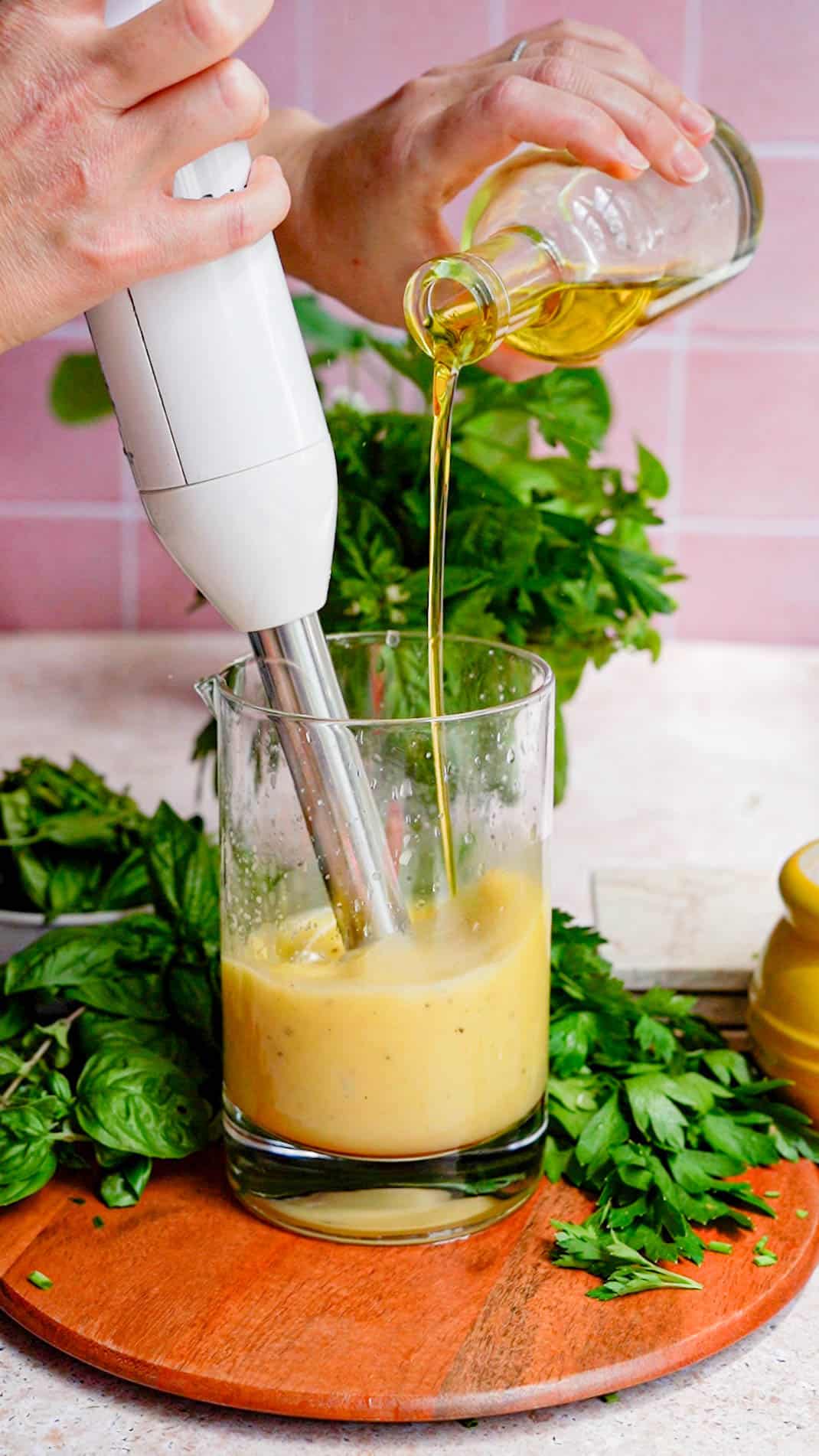 olive oil is being drizzled into a mixing jar with an immersion blender running, as a step in a recipe for a vinaigrette salad dressing.