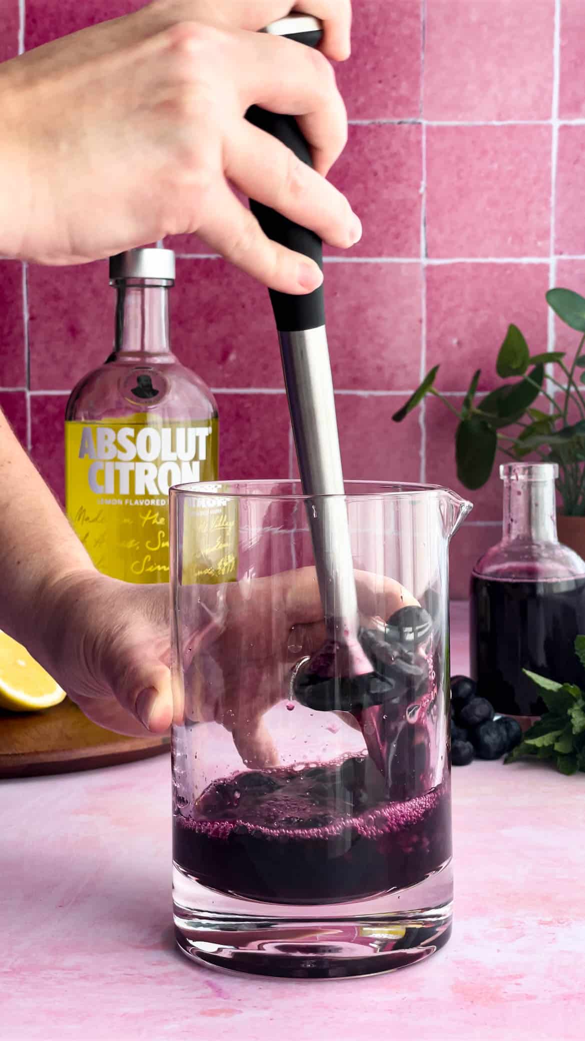 A person from out of frame is muddling blueberries, blueberry simple syrup, and lemon juice in a cocktail mixing glass while a bottle of Absolut Citron sits in the background.