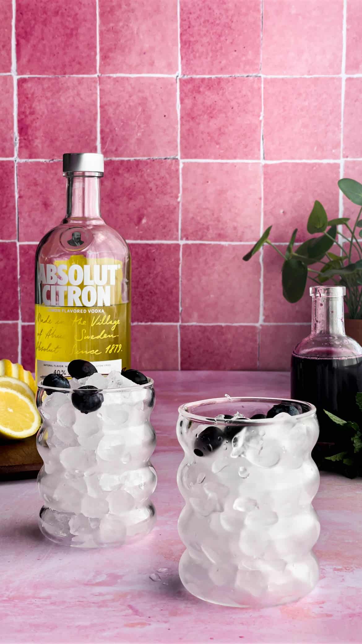 Two cocktail glasses are filled with ice and garnished with blueberries. A bottle of Absolut Citron vodka sits in the background.