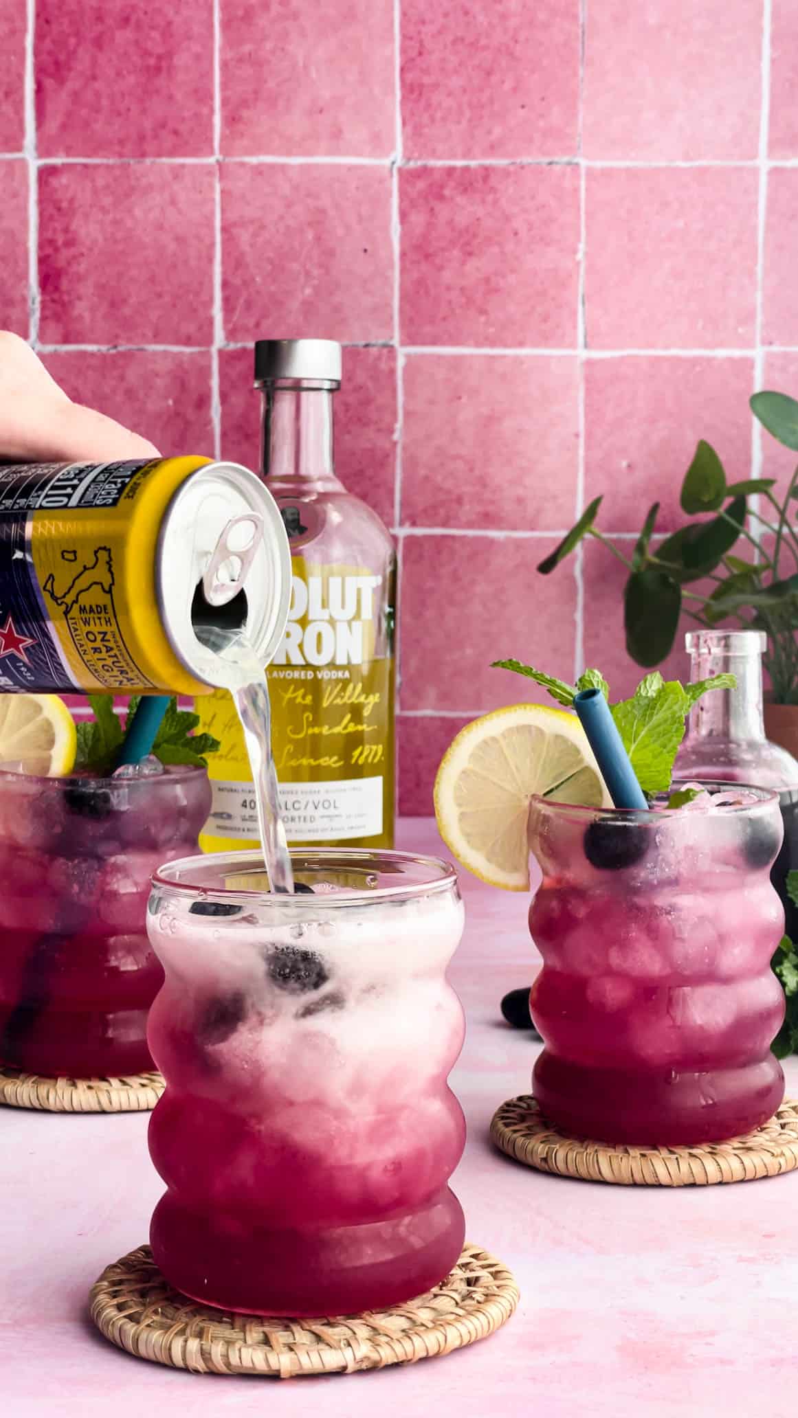 A hand from out of frame is pouring sparking lemon water into a cocktail glass to top off a sparkling blueberry vodka lemonade cocktail.