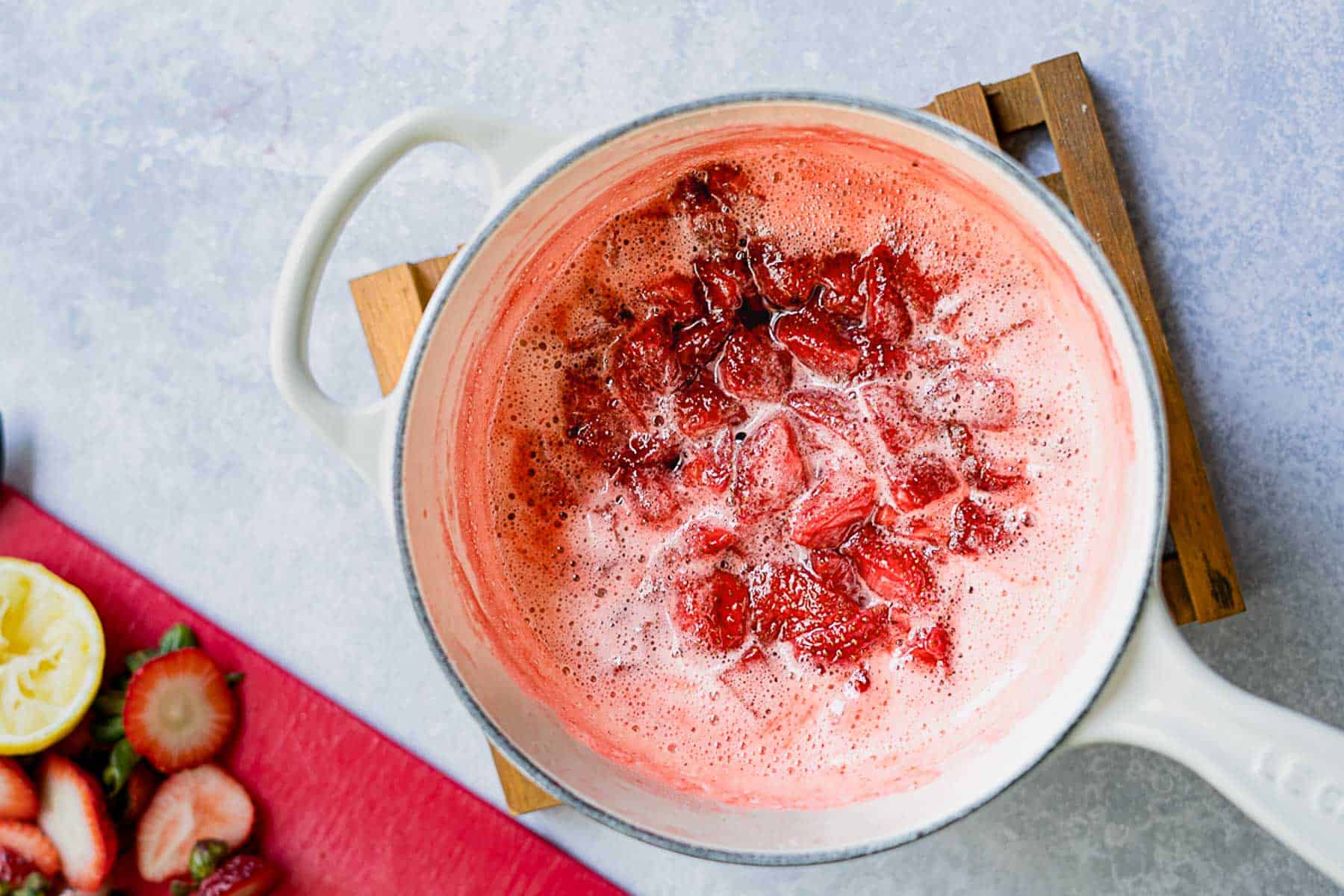 A white pot filled with bubbling chopped strawberries simmers on a wooden trivet, hinting at the creation of a delicious strawberry jam without pectin. Nearby, sliced strawberries and a squeezed lemon rest on a red cutting board, ready to add fresh flavor to the homemade preserve.