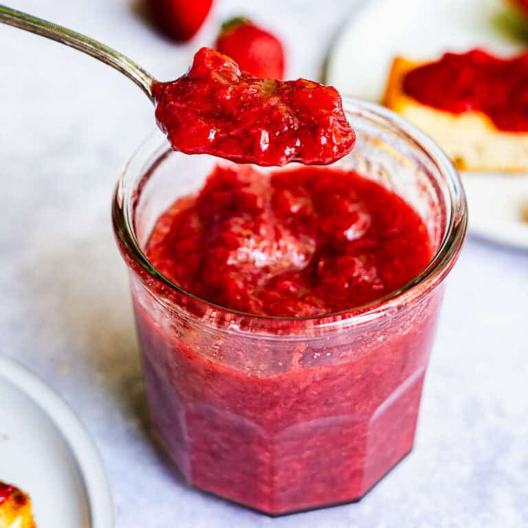A spoonful of chunky strawberry jam without pectin is held above an open glass jar filled with the same jam, with toast and strawberries blurred in the background.