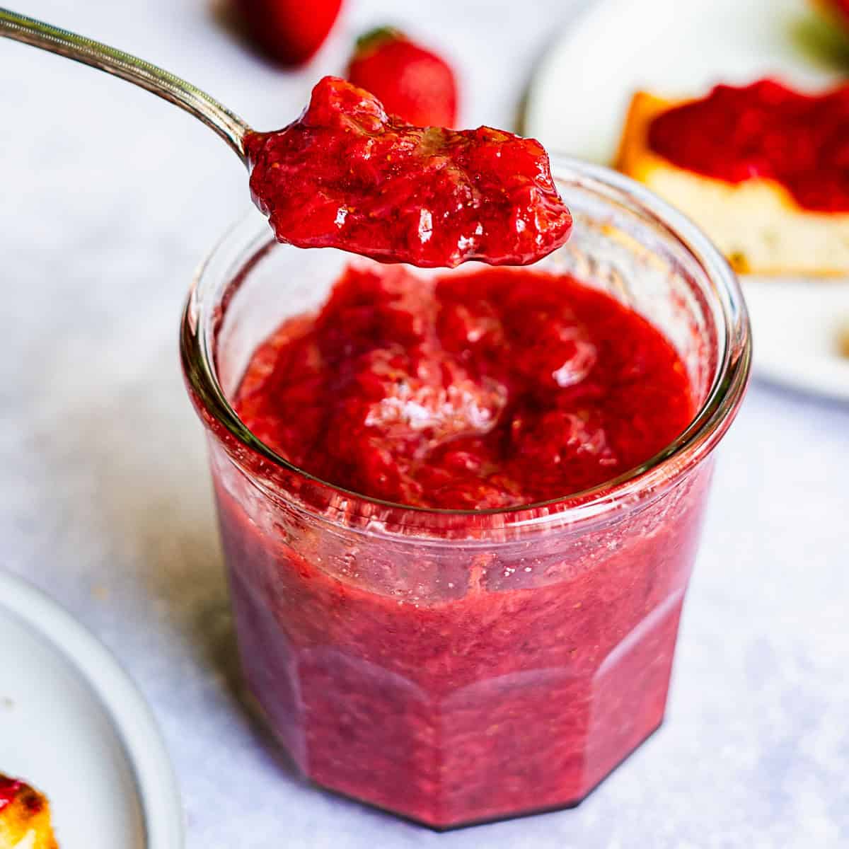 A spoonful of chunky strawberry jam without pectin is held above an open glass jar filled with the same jam, with toast and strawberries blurred in the background.