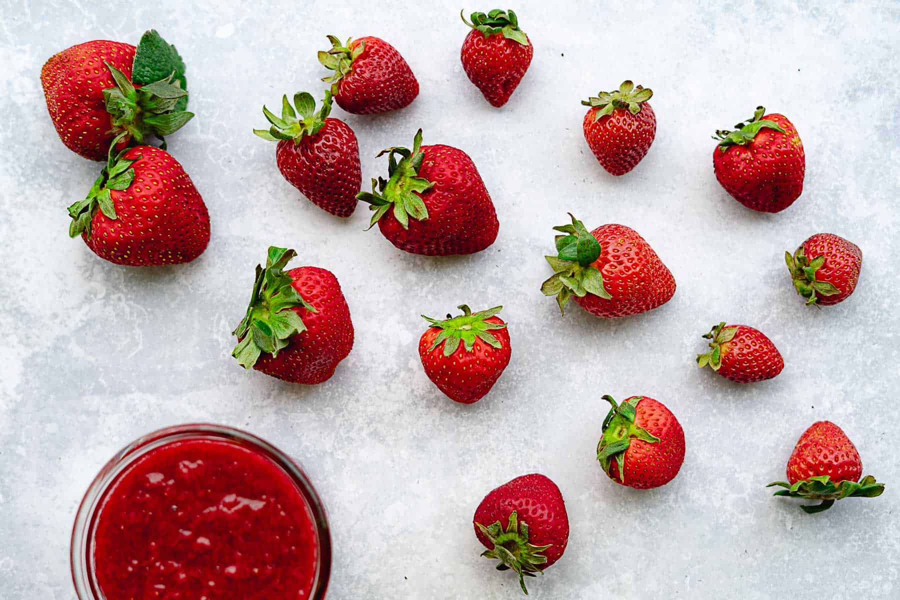 Several whole strawberries are scattered on a light surface, with a glass bowl of strawberry puree—perfect for making strawberry jam without pectin—in the lower left corner. The strawberries are fresh with green leafy tops.