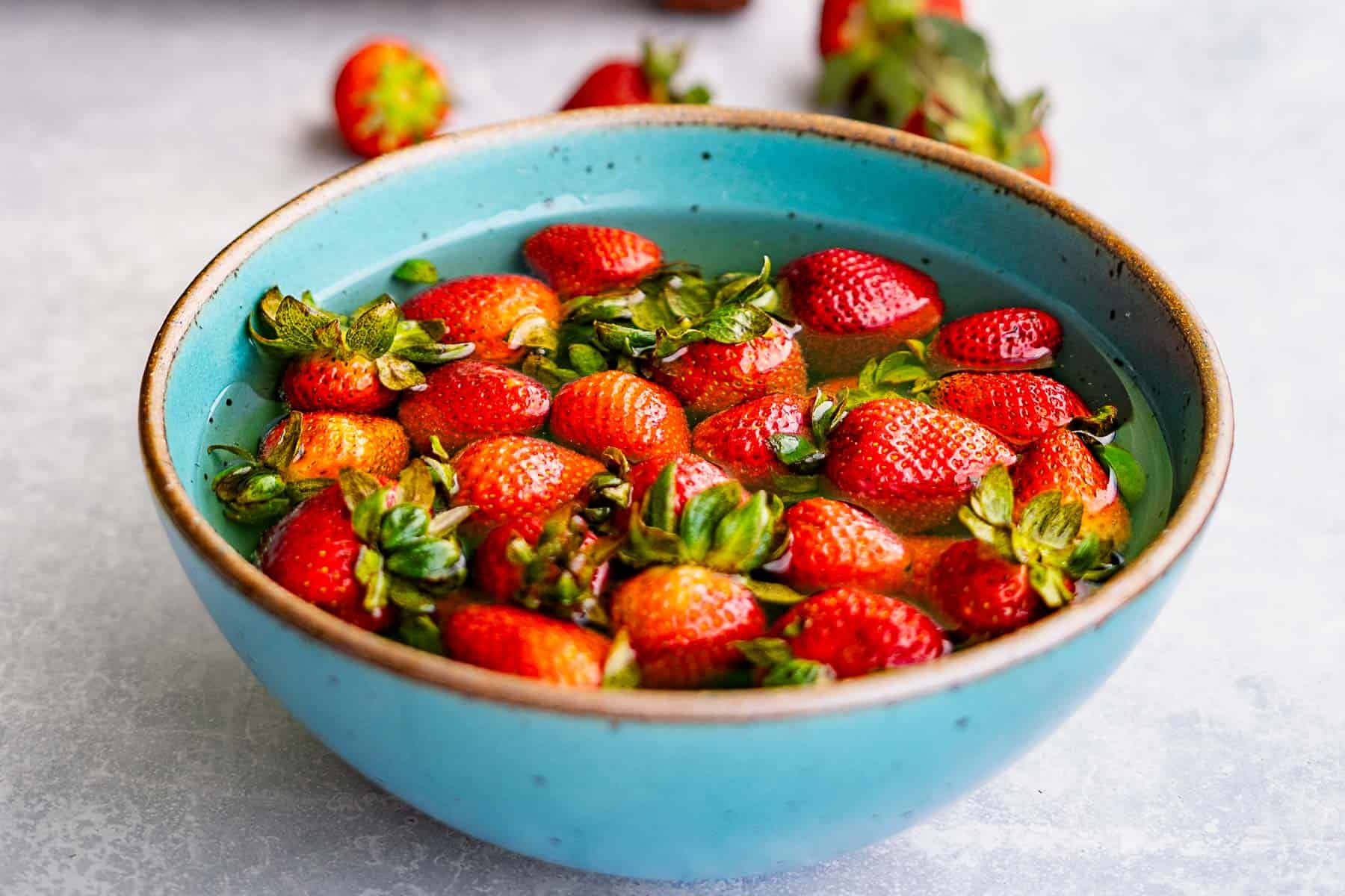 A turquoise bowl filled with fresh strawberries soaking in water, reminiscent of the perfect start for making strawberry jam without pectin, with a few strawberries in the background on a light surface.