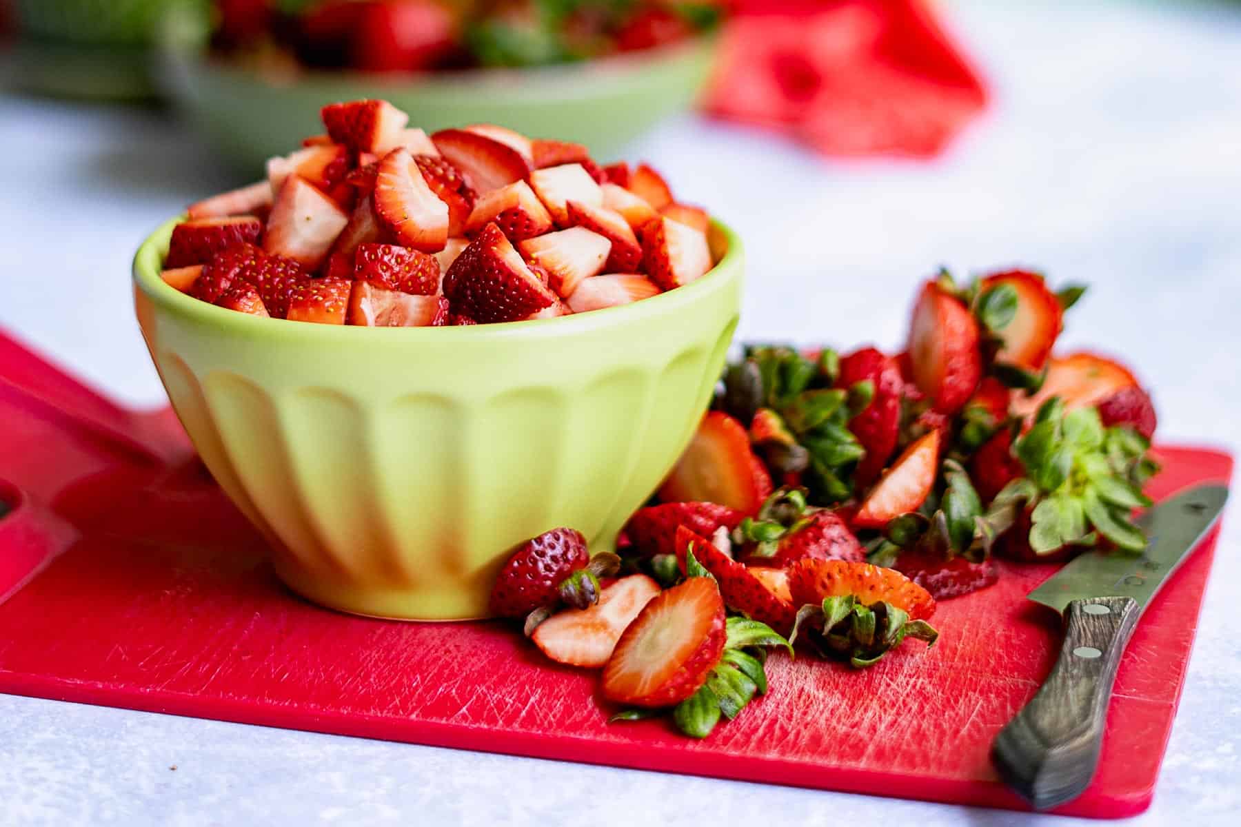 A yellow bowl filled with chopped strawberries sits on a red cutting board, surrounded by strawberry tops, slices, and a knife—perfect for preparing fresh strawberry jam without pectin.