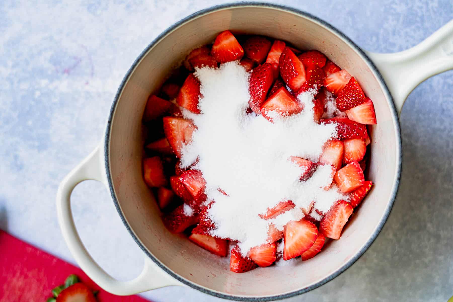A white pot filled with chopped strawberries and a mound of sugar, ready to be mixed for homemade strawberry jam without pectin, sits on a light blue surface.