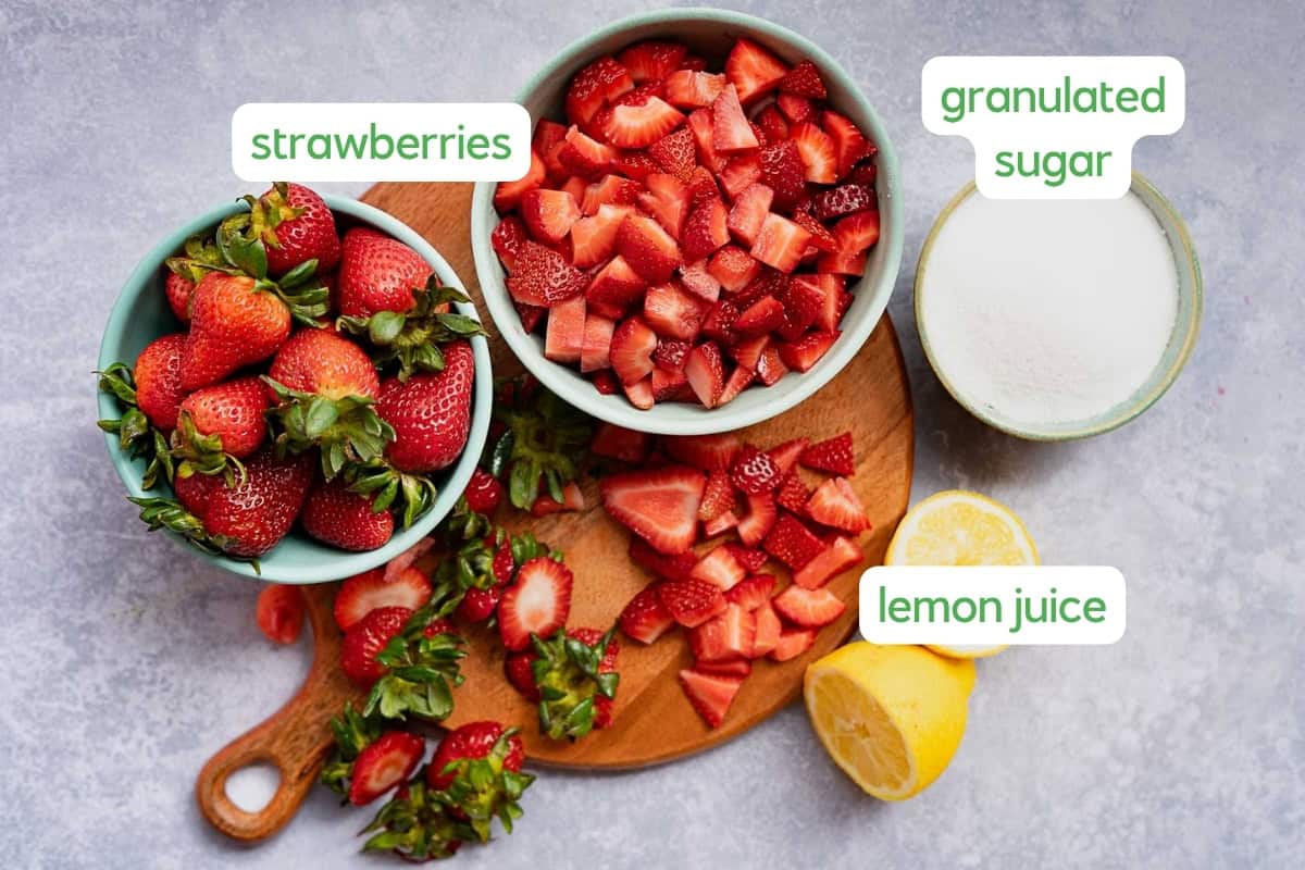 Bowls with whole and chopped strawberries, a bowl of granulated sugar, and lemon halves labeled “strawberries,” “granulated sugar,” and “lemon juice” on a wooden board—perfect for making strawberry jam without pectin.
