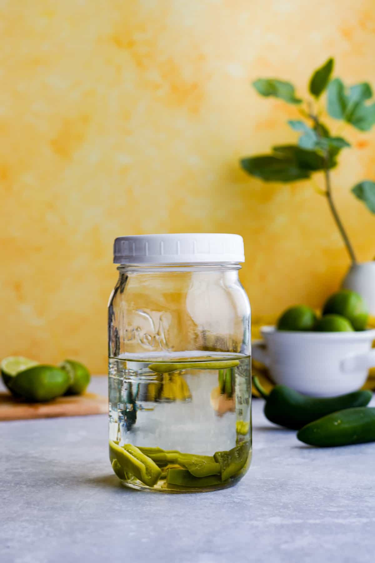 a mason jar full of jalapeño infused tequila sits on a countertop with a yellow background. The jalapeño peppers are at the base of the jar full of tequila.