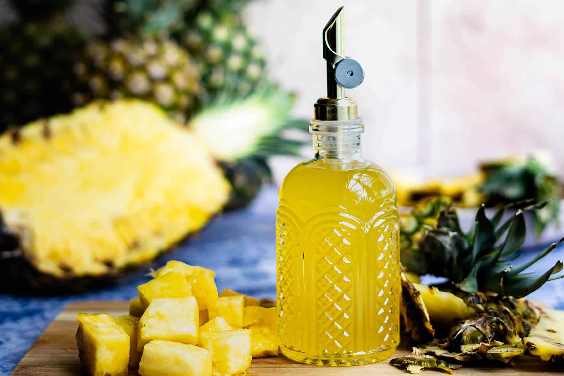 A glass bottle with a metal spout filled with yellow pineapple simple syrup stands on a wooden surface next to pineapple chunks and a halved pineapple, with more pineapple visible in the blurred background.