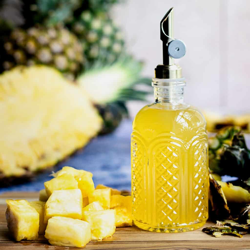 A bottle of pineapple simple syrup sits on a wooden cutting board next to a pile of pineapple chunks. There are pineapples in the background, including a sliced half of a pineapple.