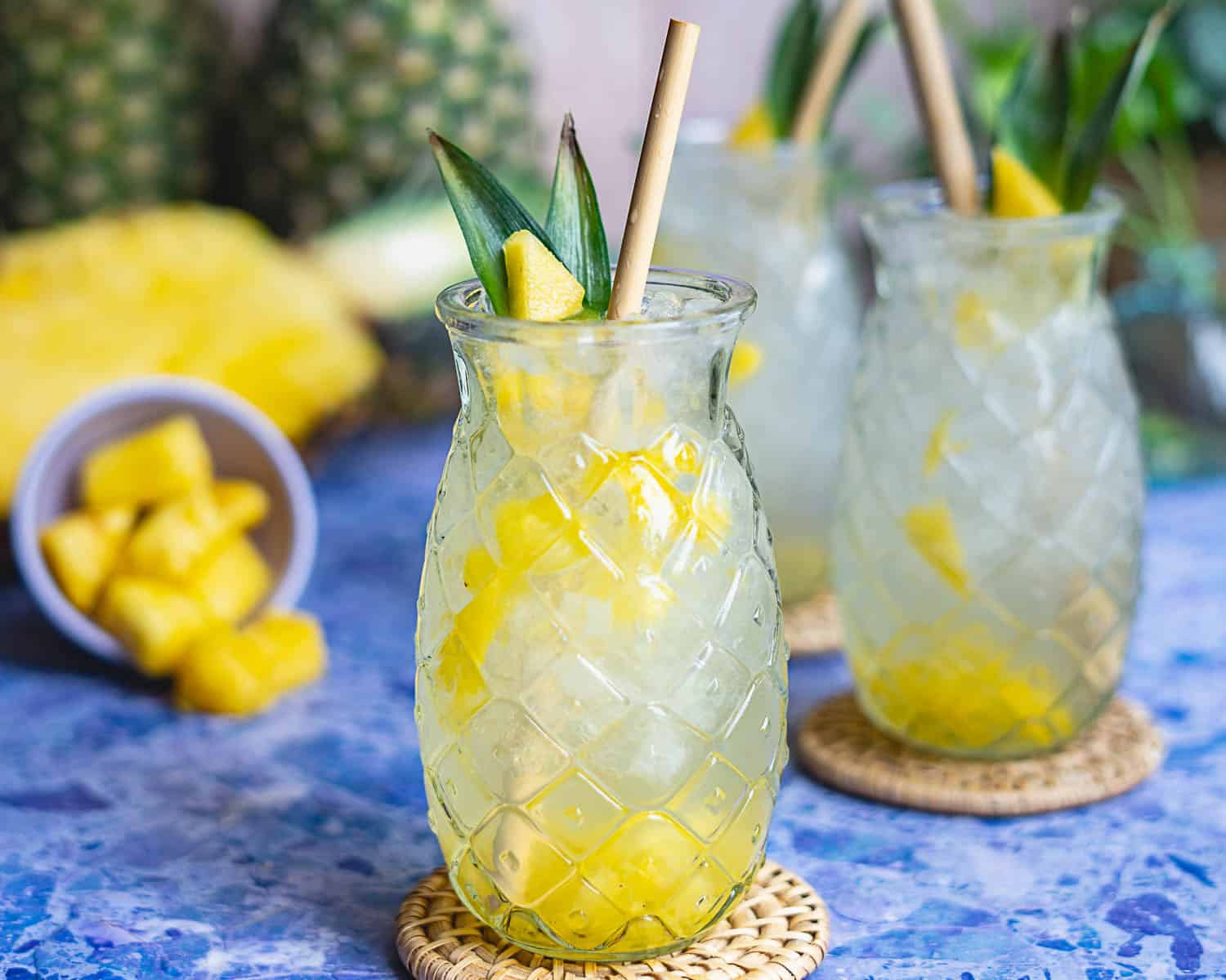 A glass with a pineapple pattern holds a tropical pineapple soda with ice and pineapple chunks, garnished with pineapple leaves and a straw. Pineapple pieces are in a small bowl beside the glass on a blue surface.