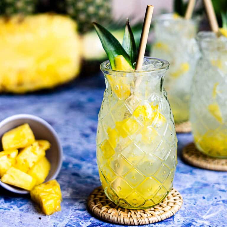 A glass with a pineapple pattern holds a tropical pineapple soda with ice and pineapple chunks, garnished with pineapple leaves and a straw. Pineapple pieces are in a small bowl beside the glass on a blue surface.