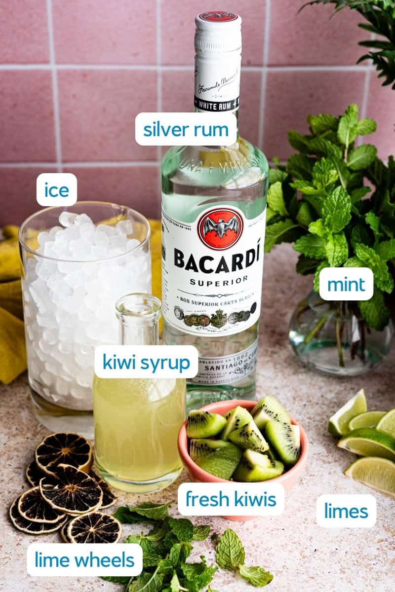 A bottle of Bacardi silver rum, a glass of ice, a jar of kiwi syrup, fresh kiwi slices, dried lime wheels, whole limes, and a bunch of mint are arranged on a counter—perfect ingredients for crafting a refreshing kiwi mojito.