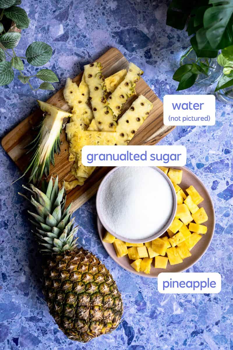 A labeled ingredient shot of the ingredients used to make pineapple simple syrup siting on a purple stone countertop.