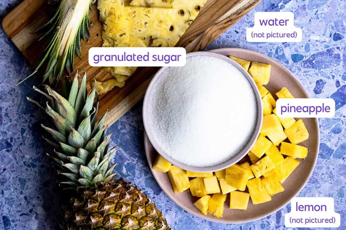 A whole pineapple, a wooden board with pineapple slices, a bowl of granulated sugar, and a plate of pineapple chunks on a blue surface—perfect ingredients for making refreshing pineapple simple syrup. Labels identify granulated sugar, pineapple, water (not pictured), and lemon (not pictured).