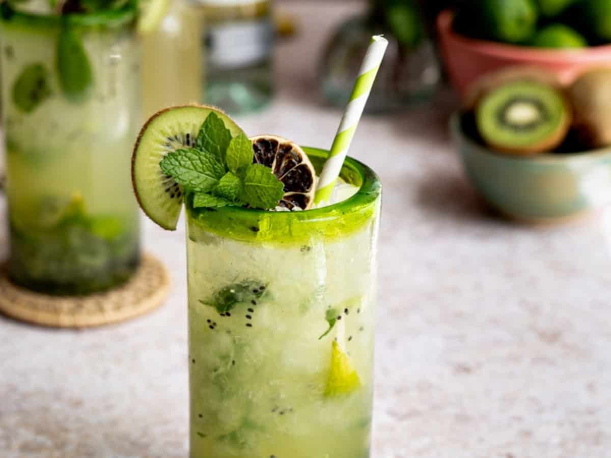 A tall glass of refreshing kiwi mojito with ice, fresh mint, lime slices, and a striped straw, garnished with a kiwi wheel. Another kiwi mojito and a bowl of fruit are blurred in the background.