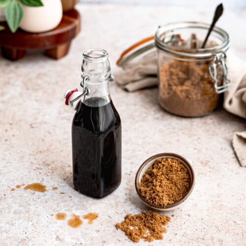 feature image of dark brown sugar simple syrup: a bottle of homemade brown sugar simple syrup is sitting on a countertop with a small dish of dark brown sugar to the right of the bottle.
