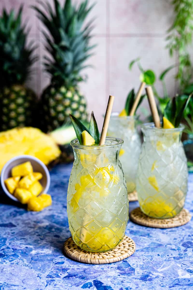 feature image of homemade pineapple soda. Three glasses of pineapple soda sit on a purple stone countertop, with cubes of pineapple to the left and a half pineapple in the background.