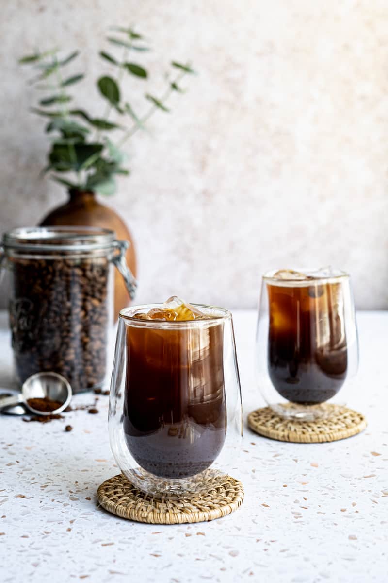 Feature image of an iced Americano coffee beverage. Two iced Americanos sit on a terrazzo countertop, with a container of coffee beans in the background.