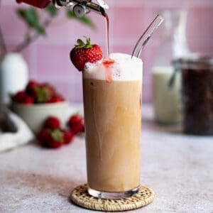 Feature image of a iced strawberry latte. A bottle from out of frame drizzles strawberry simple syrup onto an iced strawberry latte.