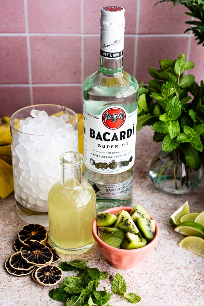 Ingredients used to make kiwi mojitos sit on a light brown countertop. Ingredients include rum, ice, limes, mint leaves, kiwi fruit, and kiwi simple syrup.
